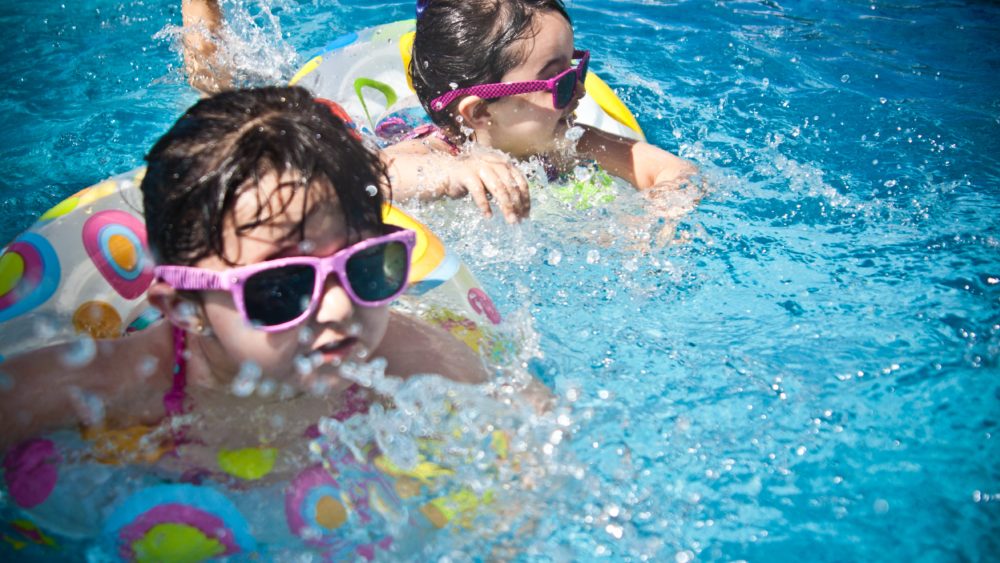 2 young girls swimming. Both are in floating rings and wearing sunglasses.