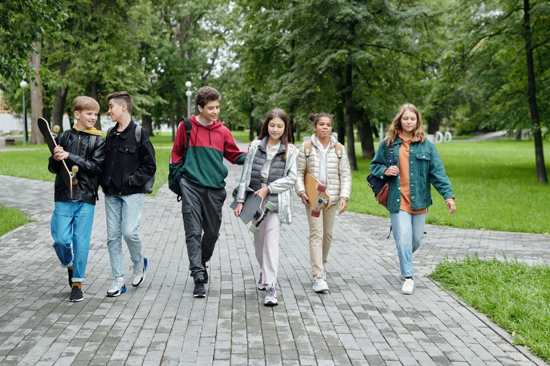 Group of teenaged girls and boys walking in a park with backpacks and skateboards.